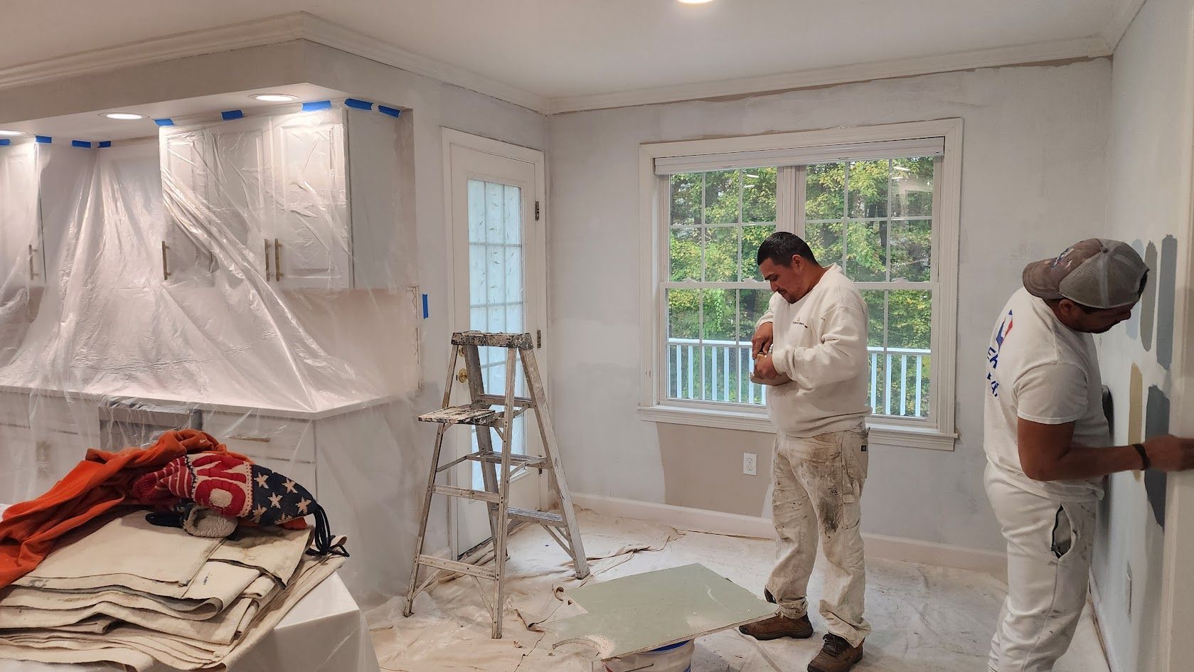 Workers painting a bright white room, with plastic covering cabinets and furniture during renovation.