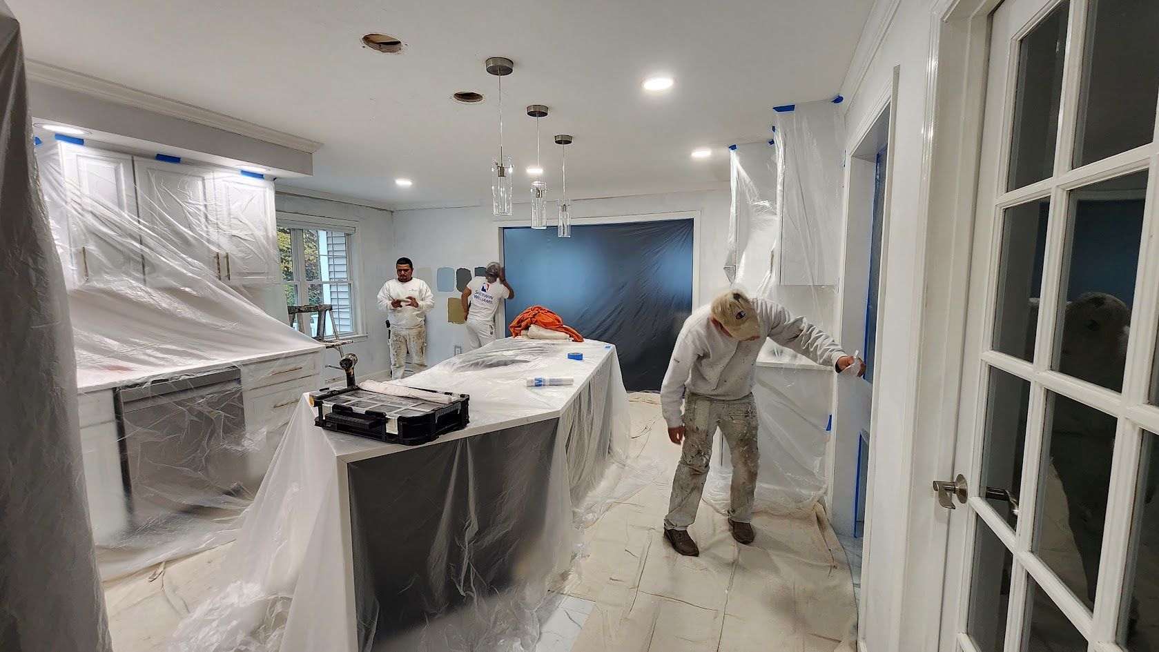 Workers masking and painting a bright hallway, with plastic-covered furniture and walls.