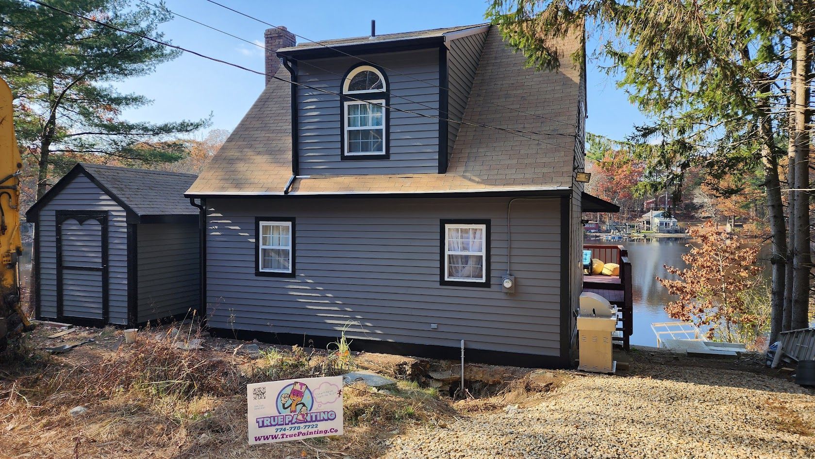 Gray house with dormer and shed, autumn trees, and a small sign in the front yard.