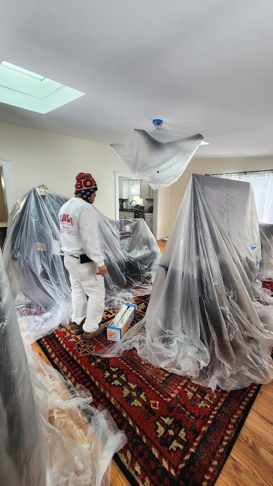 Workers in white hazmat suits spray and cover a room with plastic sheeting over rugs and furniture.