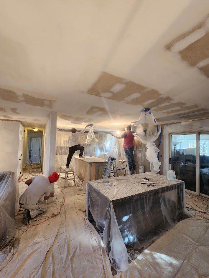 Workers mudding and taping a room during drywall renovation, with plastic-covered furniture and ceiling patches.