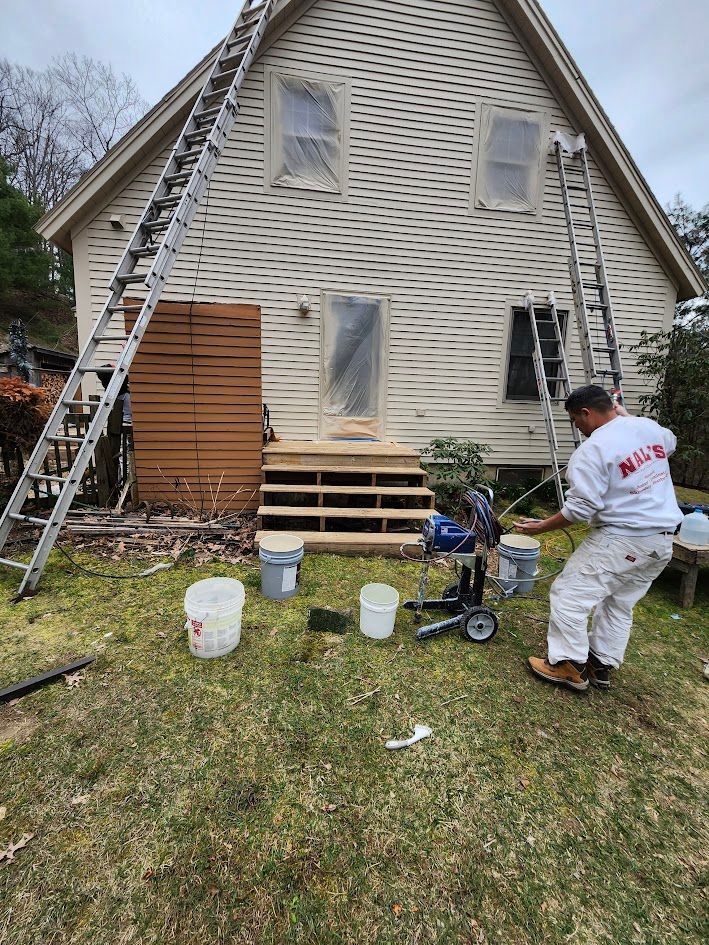Workers spray-painting the front of a beige A-frame house with ladders and buckets on the lawn