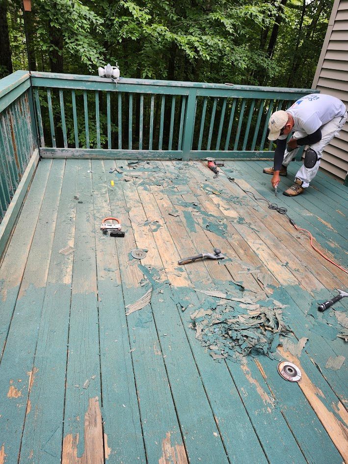 Worker scraping peeling paint from a weathered green wooden deck on a porch.