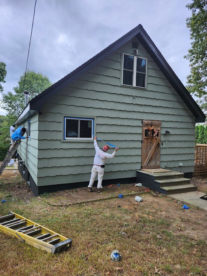 Two workers painting the exterior of a small gray house with ladders and tools on the lawn
