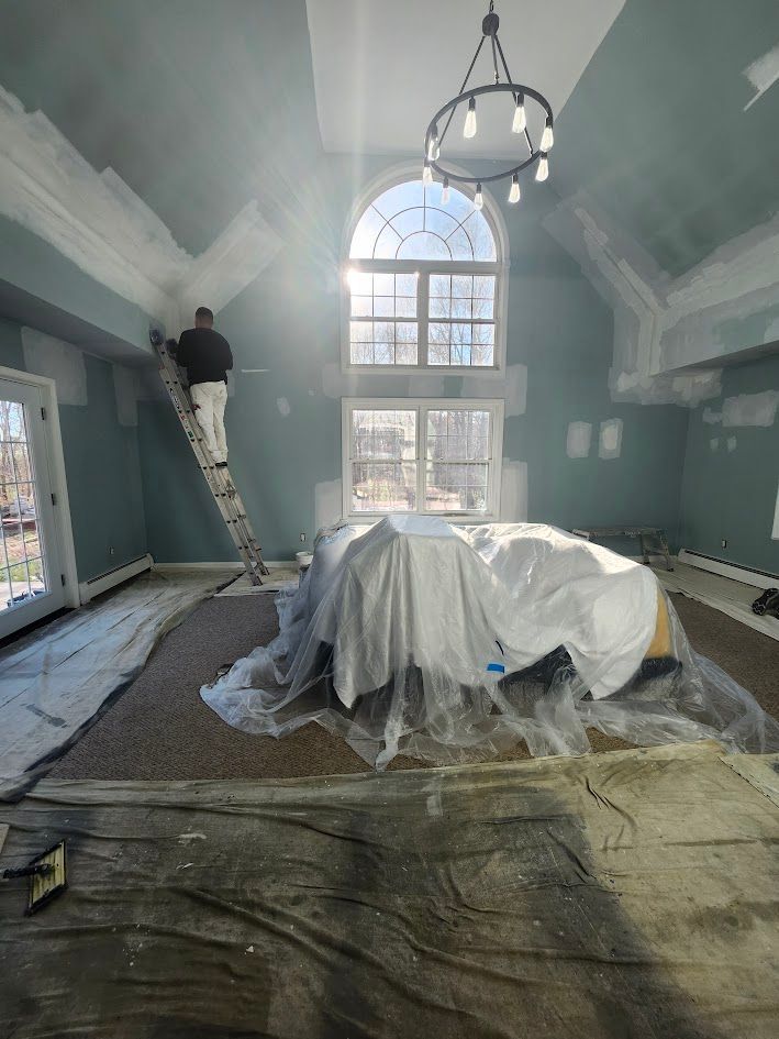 Renovated room with vaulted ceiling, chandelier, covered furniture, and worker painting the upper wall.