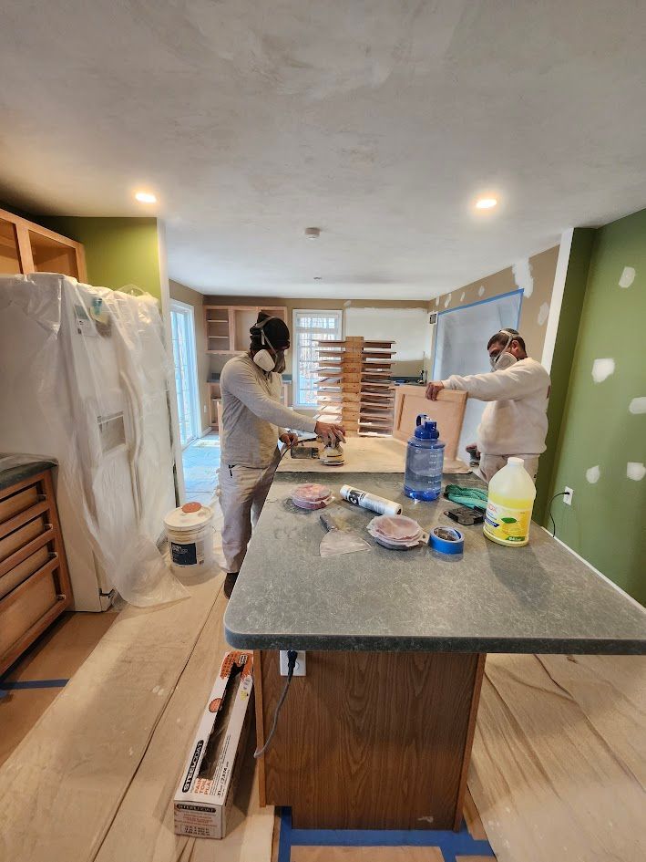 Two people in a kitchen appear to be painting walls around a central island and appliances.