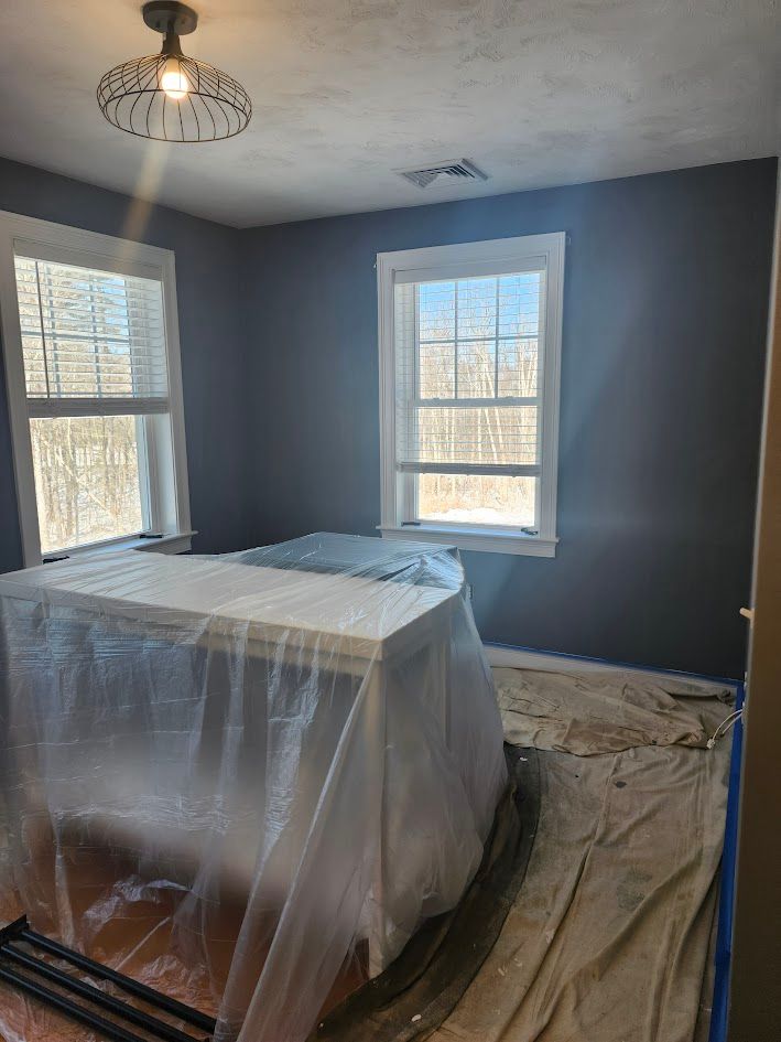 Small room being painted gray, with plastic-covered furniture and windows letting in daylight.