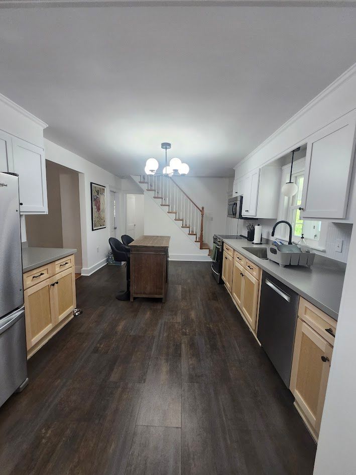 Bright kitchen with dark wood floor, white cabinets, gray countertops, and a staircase at the back.