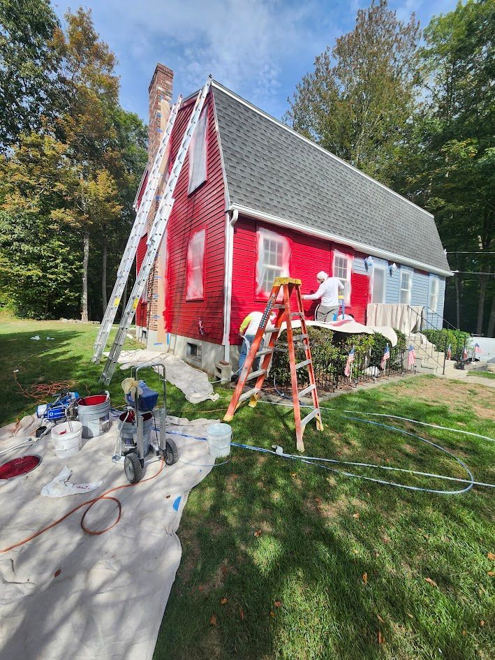 Person painting the exterior of a red house with a ladder and supplies in a sunny yard