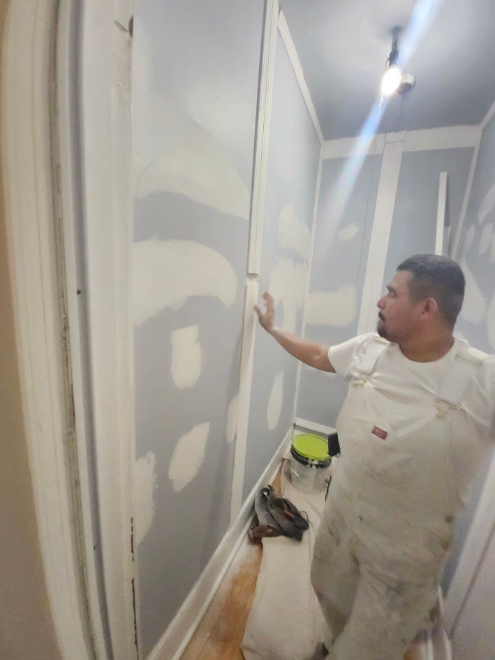 Painter smoothing gray drywall in a narrow room with patchwork seams and a bucket on the floor