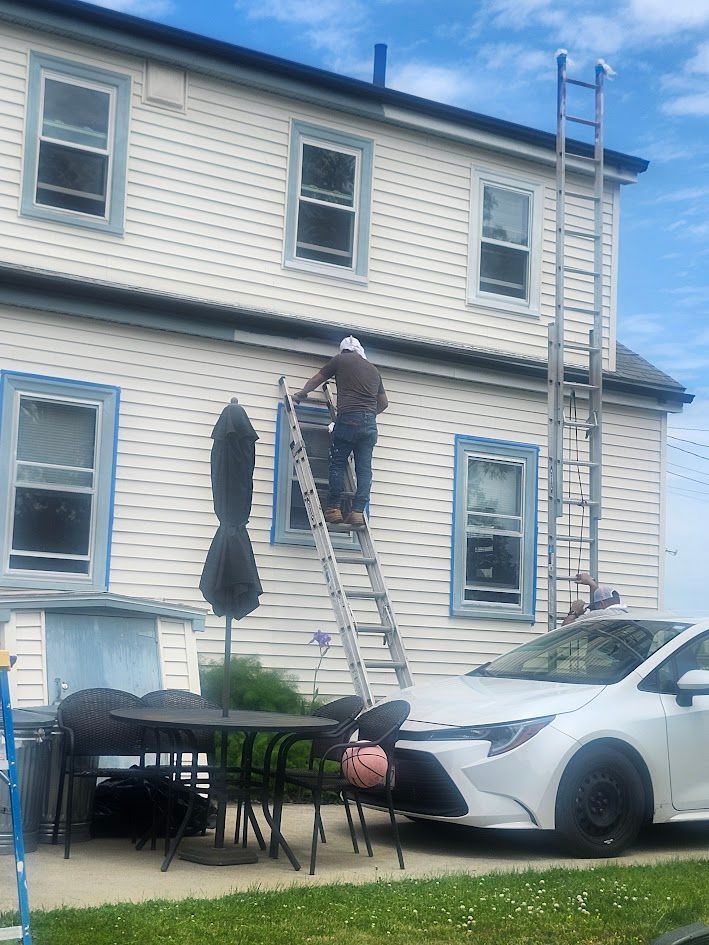 Worker on a ladder painting a white two-story house beside a parked car.