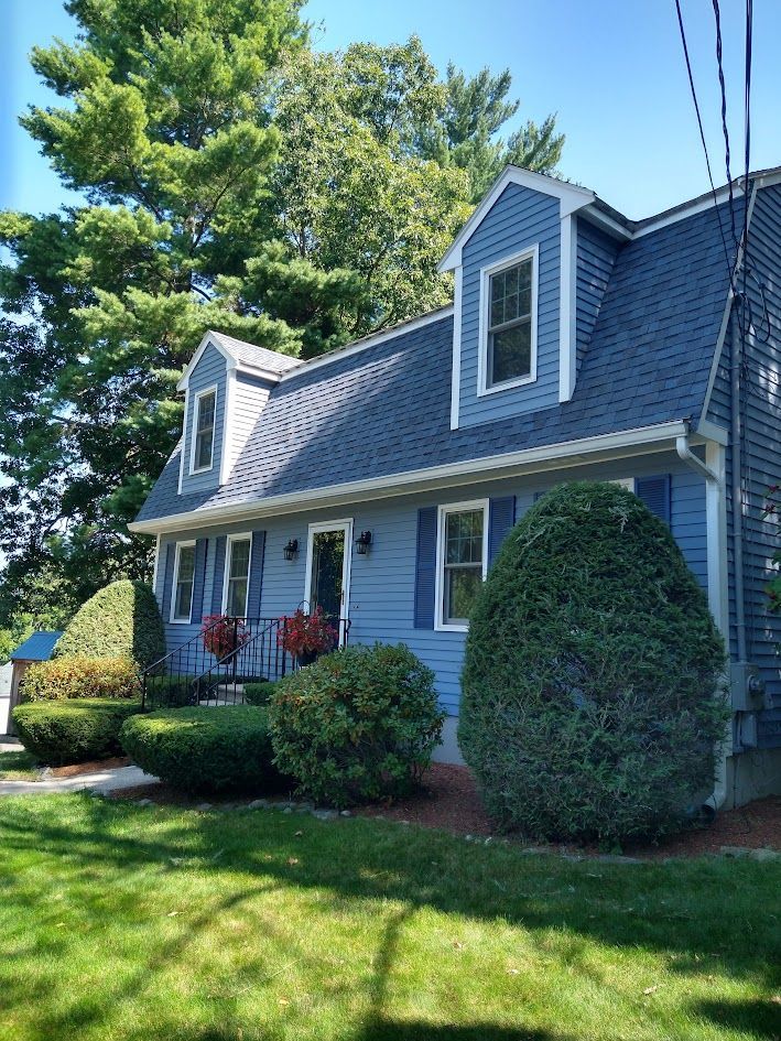 Gray house with blue shutters and dormer windows, front porch, manicured shrubs, and green lawn under sunny sky
