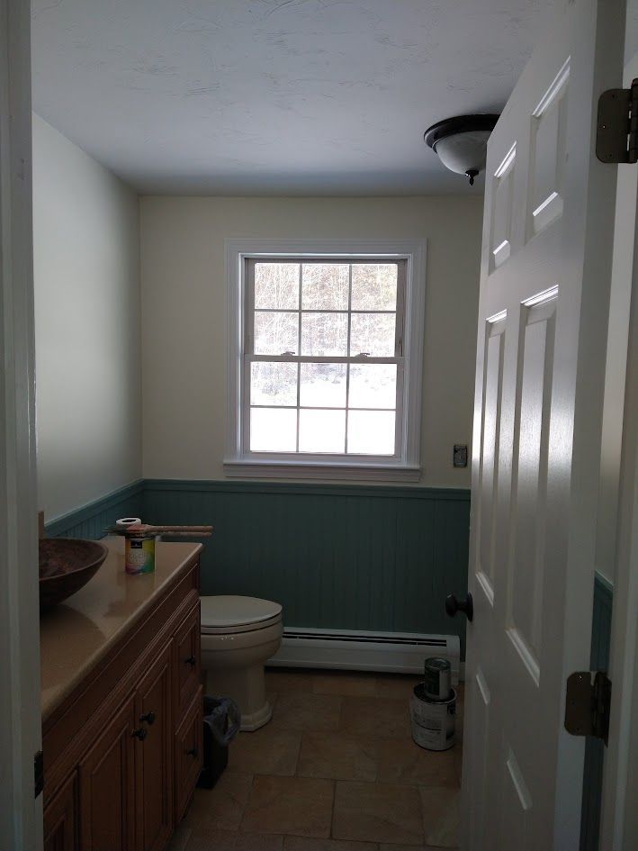 Small bathroom with white walls, green wainscoting, a window, toilet, sink cabinet, and open door.