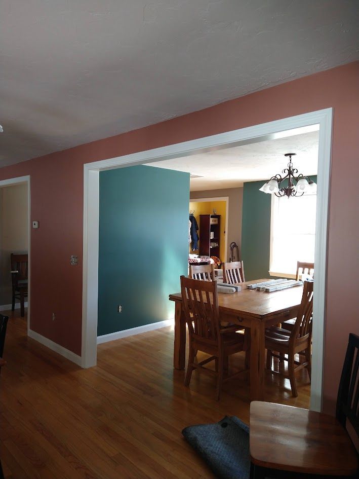 Dining room with wooden table and chairs, teal wall, and doorway opening to a bright kitchen.