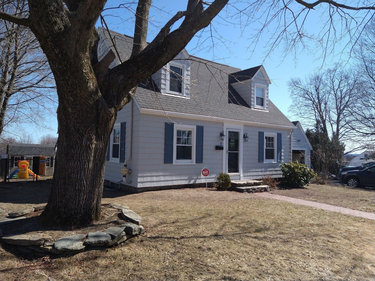 White two-story house with blue shutters, a large tree in front, and a driveway on a sunny day