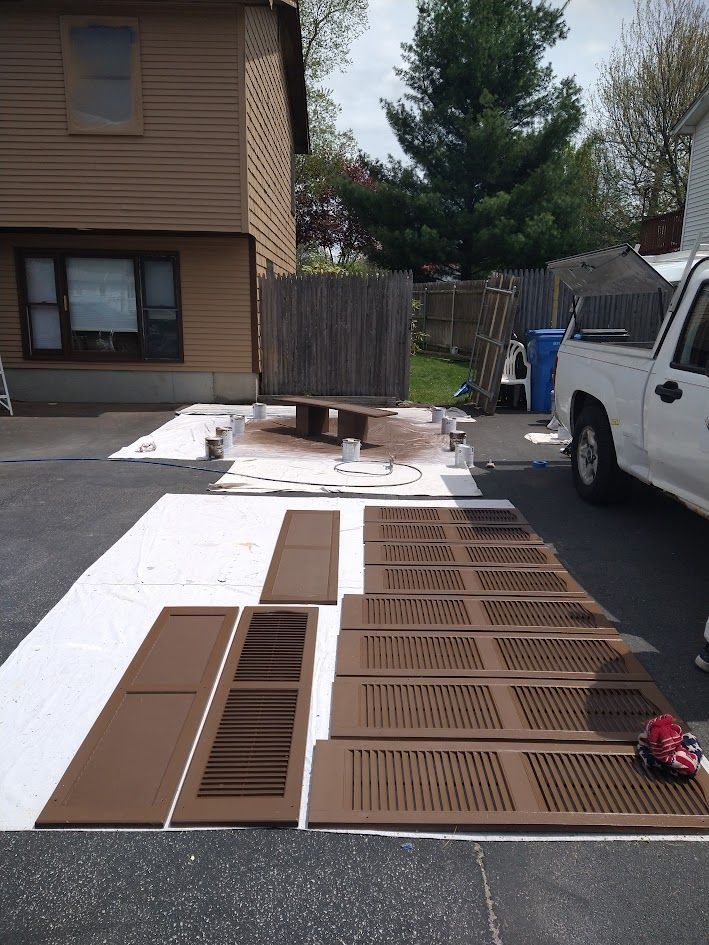 Brown window shutters laid out on white tarp in a driveway beside a house and white van.