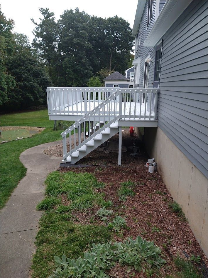 White backyard deck with stairs beside gray house, next to a curved sidewalk and landscaped lawn.