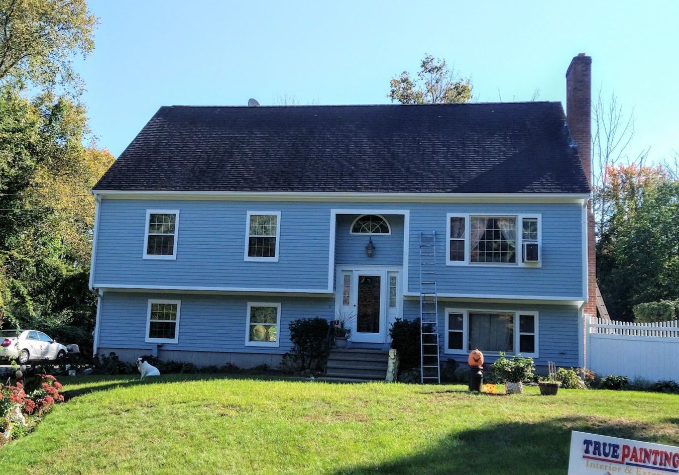 Blue two-story house with white trim, black roof, front steps, and a green lawn on a sunny day