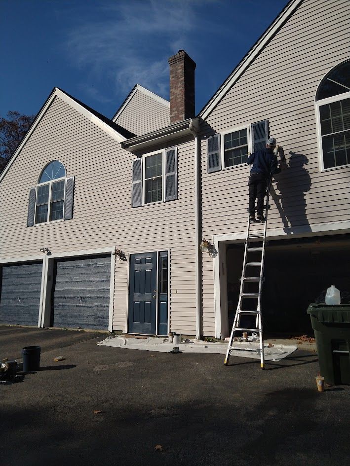 Worker on a ladder painting the upper exterior of a beige townhouse with blue garage doors.