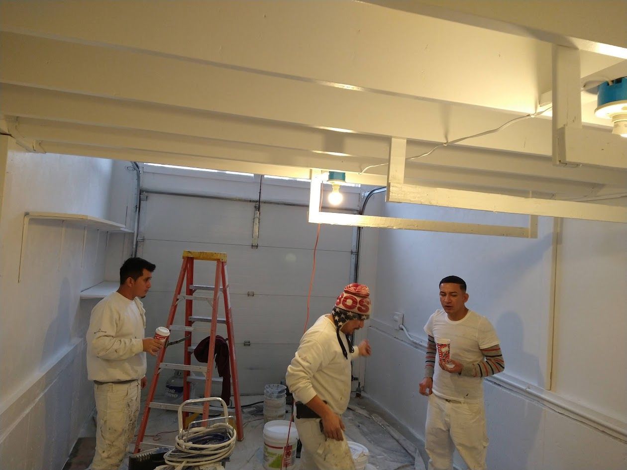 Three workers in white coveralls in a basement, one on a ladder amid renovation tools.