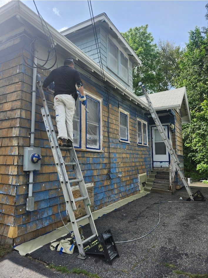 Person painting a blue-and-brick house from a ladder on the side of the driveway.