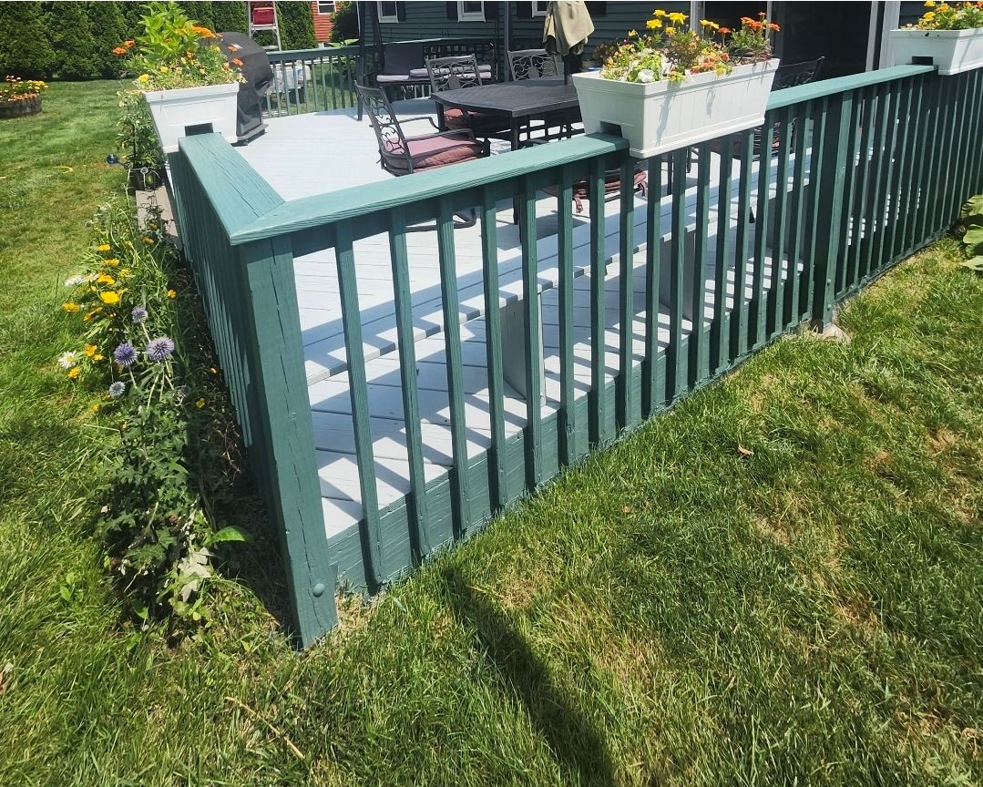 Green wooden deck railing with steps leading to a patio in a sunlit backyard garden