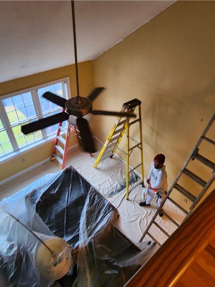 Person standing on a ladder in a room with a large ceiling fan and covered furniture during painting work