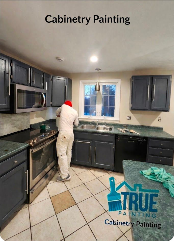 Painter in white gear painting dark kitchen cabinets in a tiled kitchen under the “Cabinetry Painting” text.