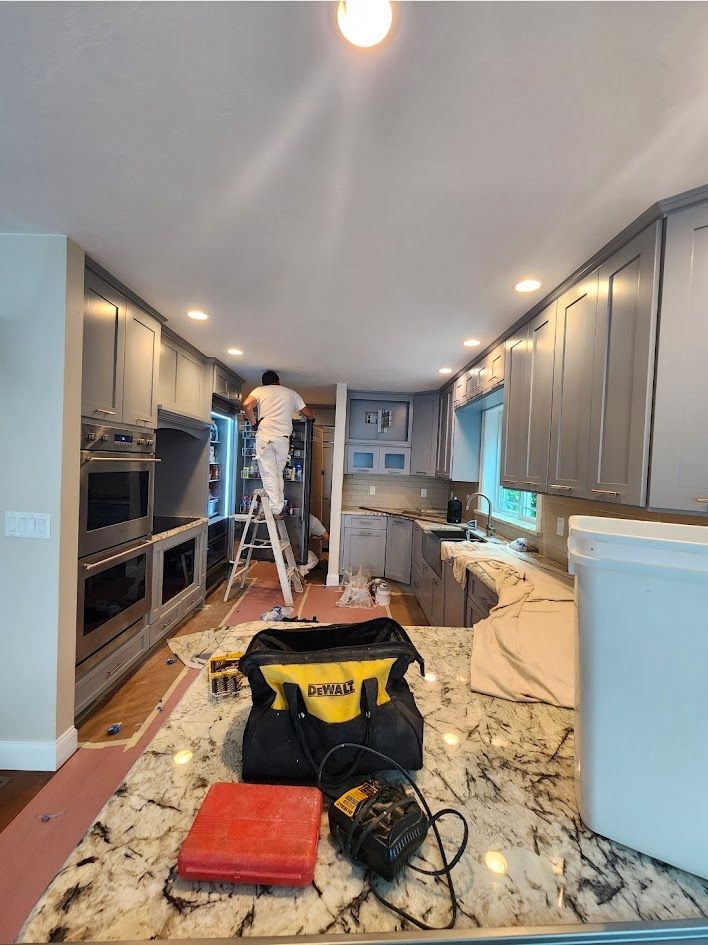Kitchen renovation in progress with cabinets installed, a worker on a ladder, and tools on a covered floor.