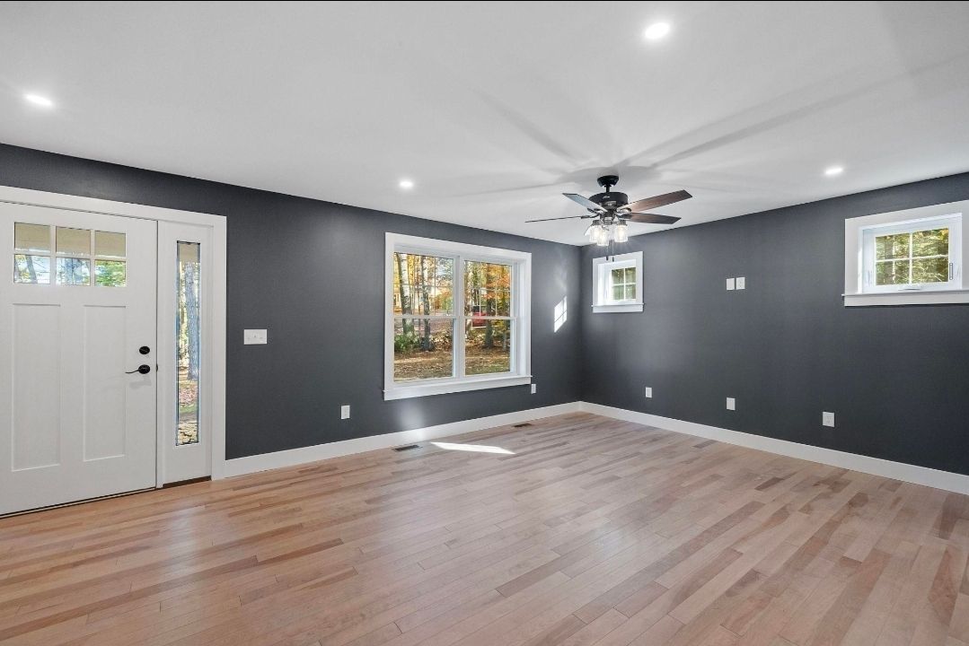 Empty living room with gray walls, light wood floors, white door, windows, and ceiling fan