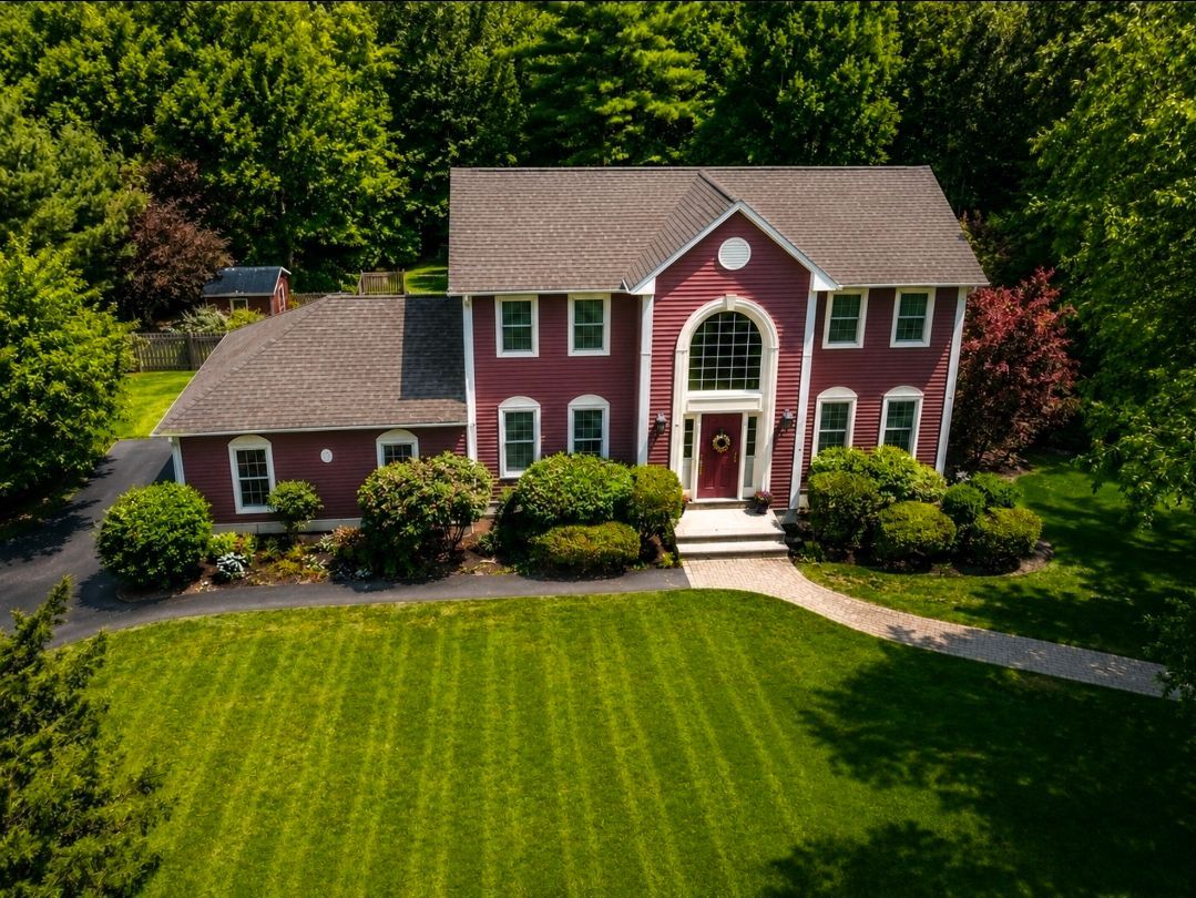 Red brick two-story house with white trim, manicured lawn, and a curved walkway surrounded by trees