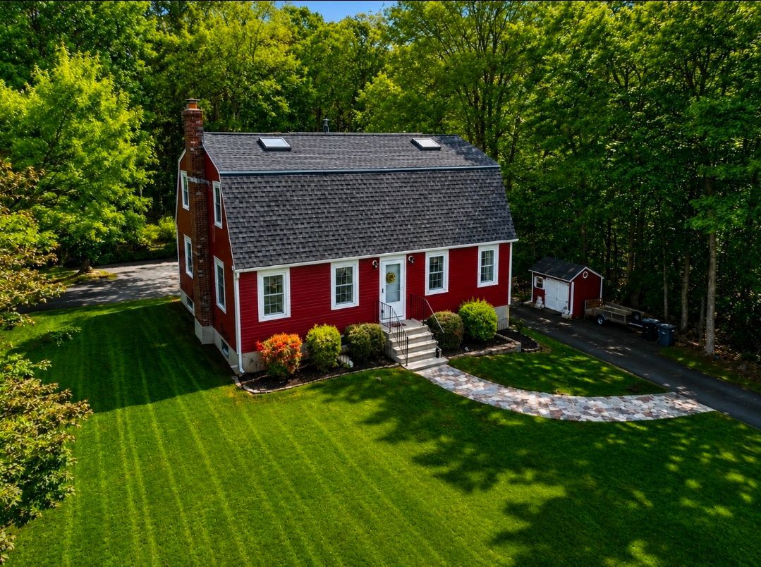 Red house with gray roof and white trim on a green lawn, surrounded by trees