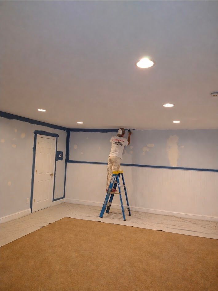 Person painting a room with blue tape, standing on a ladder in a mostly empty carpeted interior