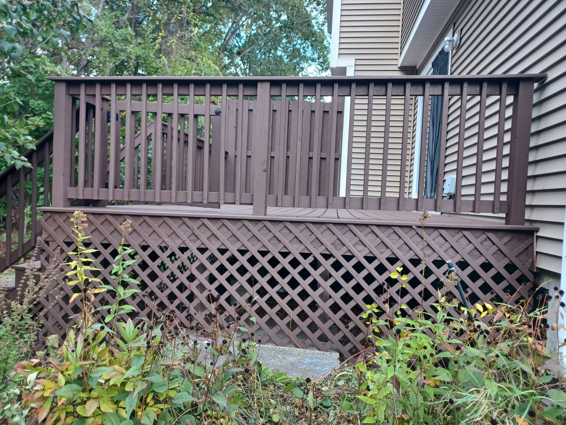 Brown wooden deck with lattice skirting, railing, and surrounding greenery beside a house