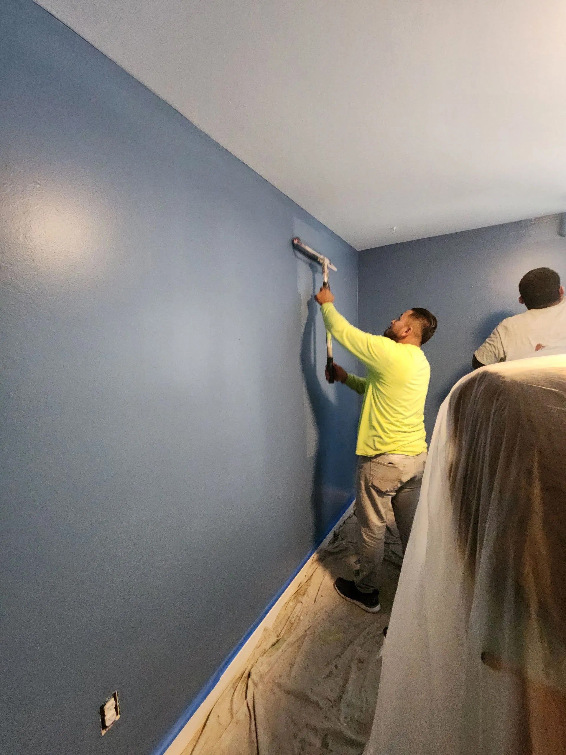 Two people painting a blue room, one rolling paint near the ceiling beside taped walls and drop cloths.