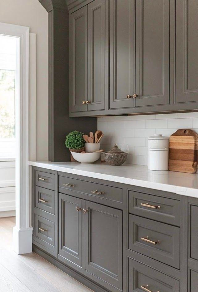 Gray kitchen cabinets with brass handles, white countertop, and small decor near a bright window