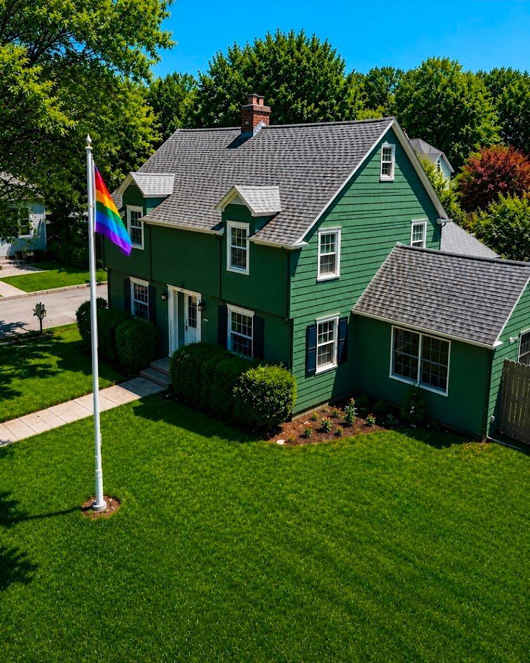 Green house with white trim and a rainbow flag on the lawn under a sunny blue sky.