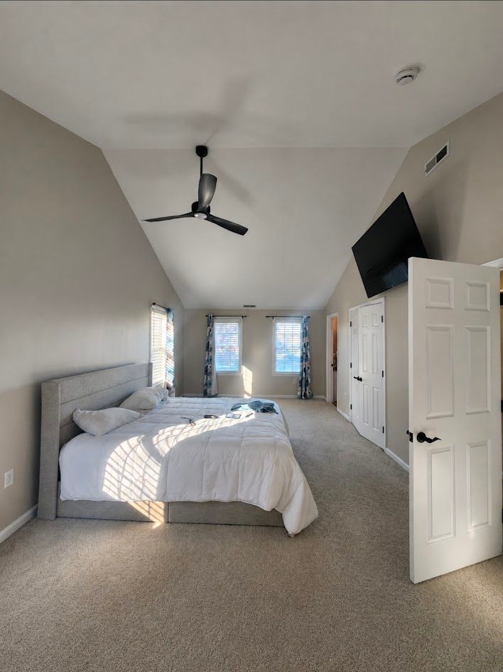 Bright bedroom with a gray bed, ceiling fan, wall-mounted TV, open door, and windows at the far end.