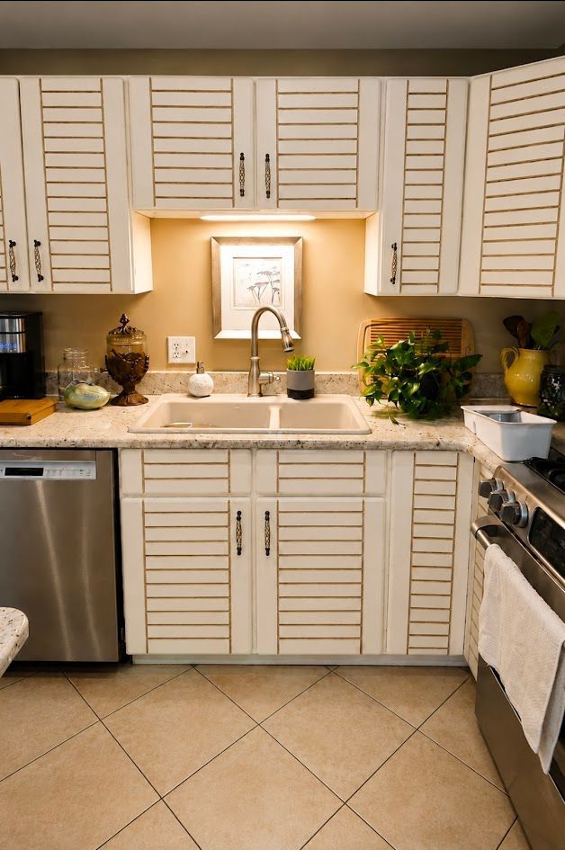 White kitchen with tan tile floor, cream cabinets, sink, dishwasher, and potted plants by the window.