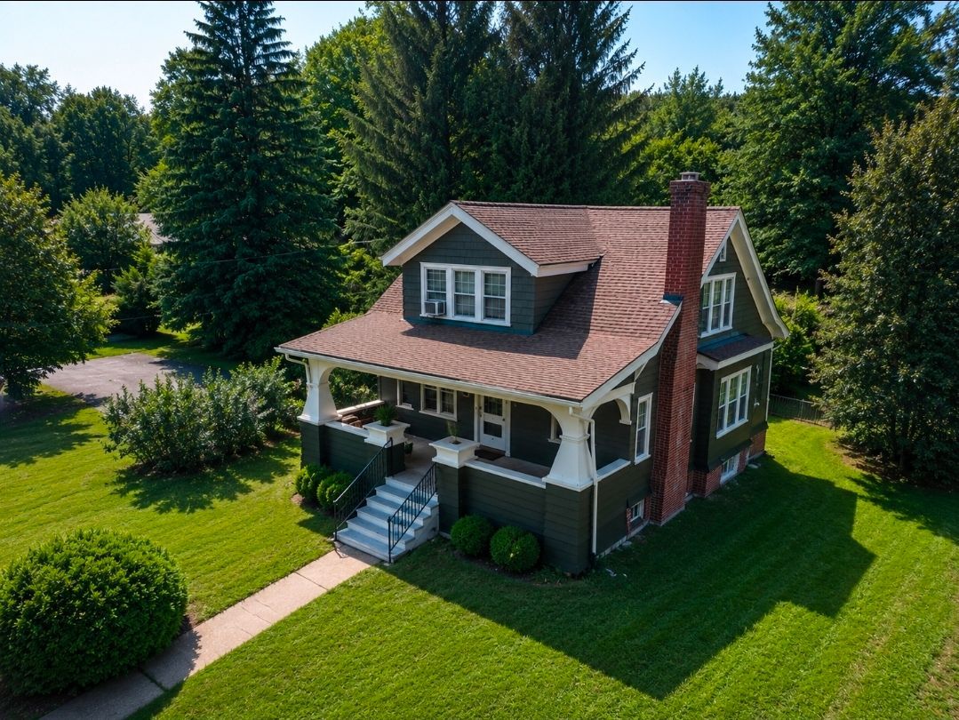 Aerial view of a small red-roofed house with a porch, surrounded by a lush green lawn and tall trees.