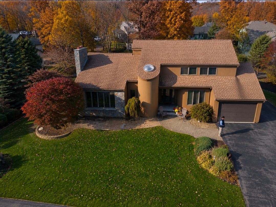 Aerial view of a suburban house with tan roof, green lawn, and autumn trees.