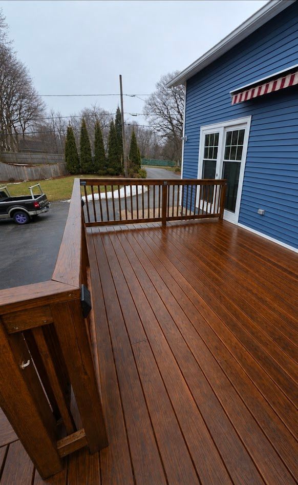 Wooden deck with railing beside a blue house, overlooking a wet driveway and yard.