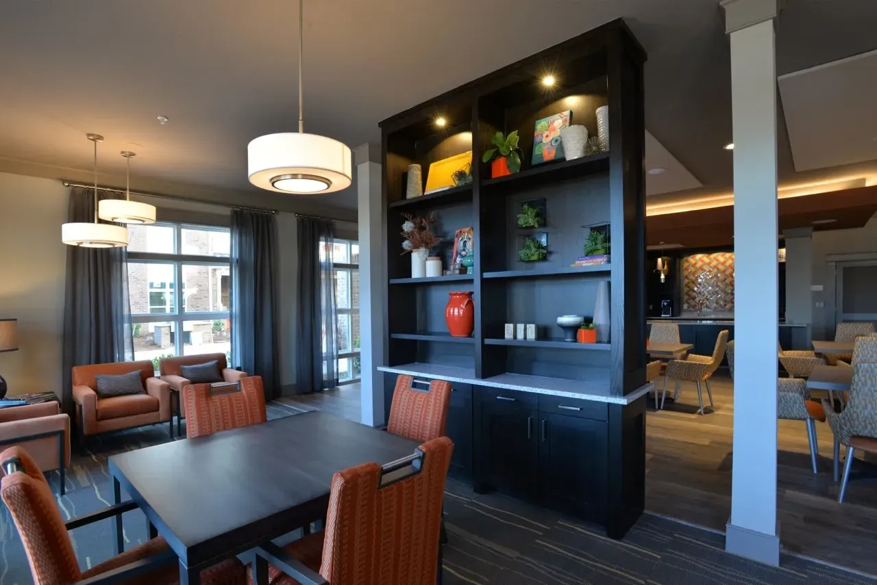 Spacious community lounge with orange upholstered chairs, dark shelving unit, and large windows.