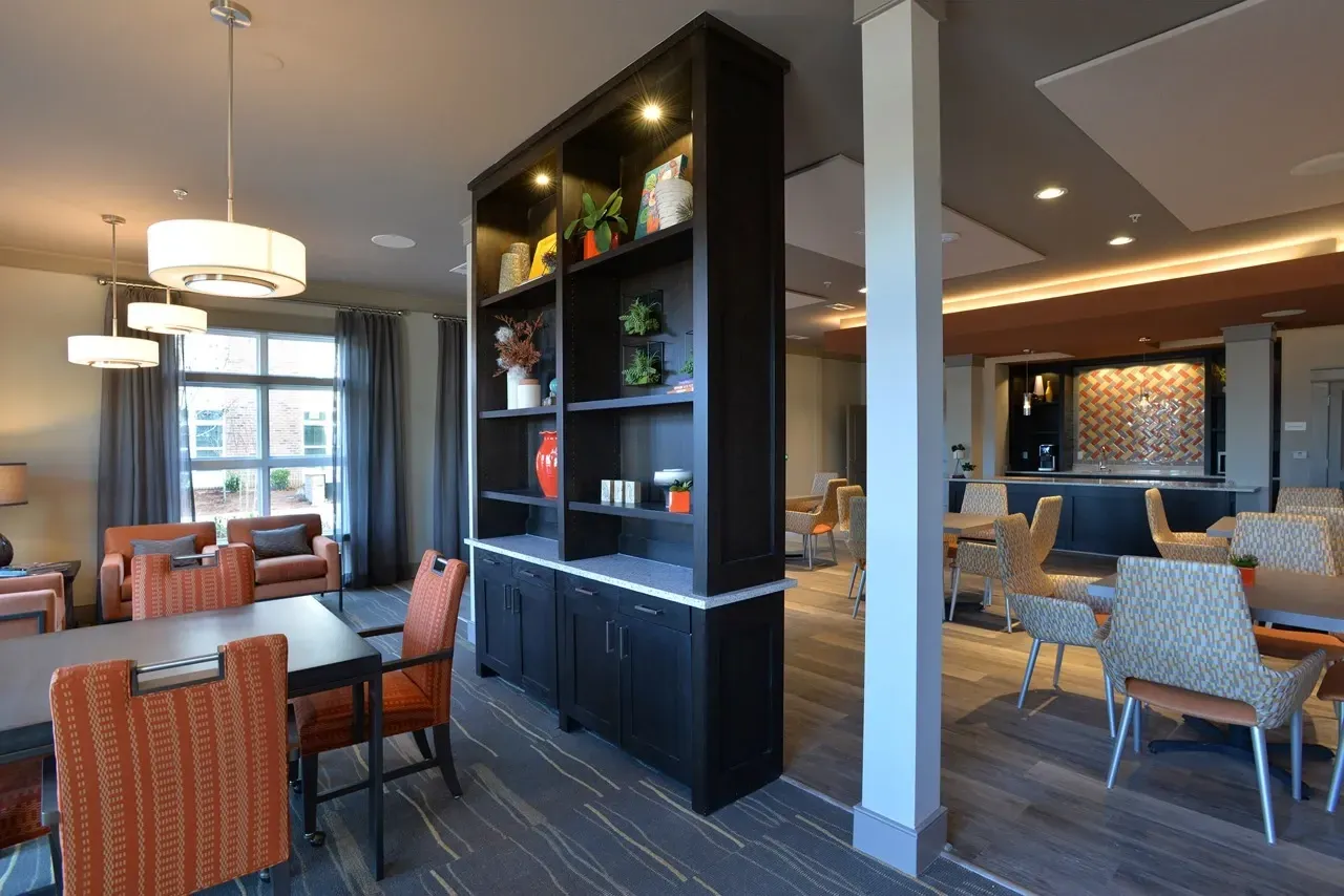 Interior of a modern apartment community lounge with seating, tables, and a tall dark shelving unit.