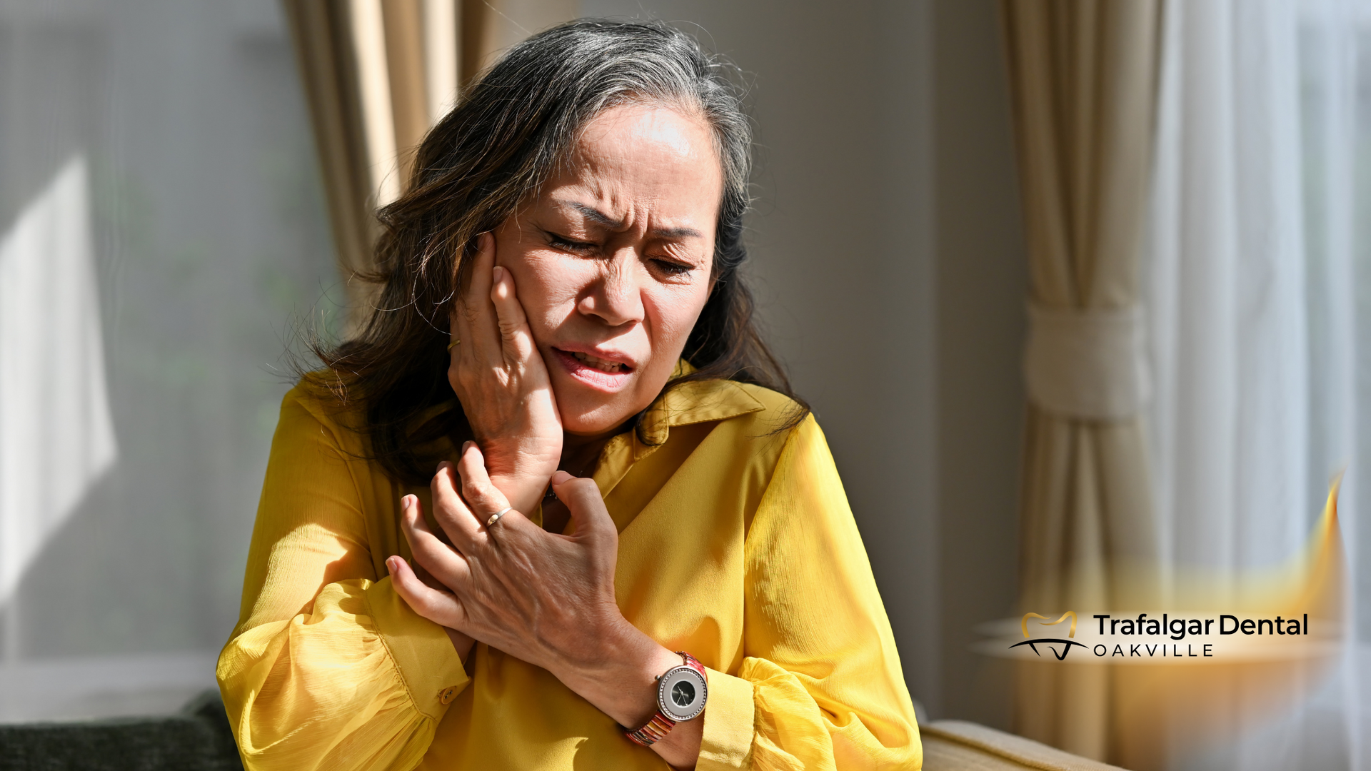 A woman in a yellow shirt is holding her face because she has a toothache.