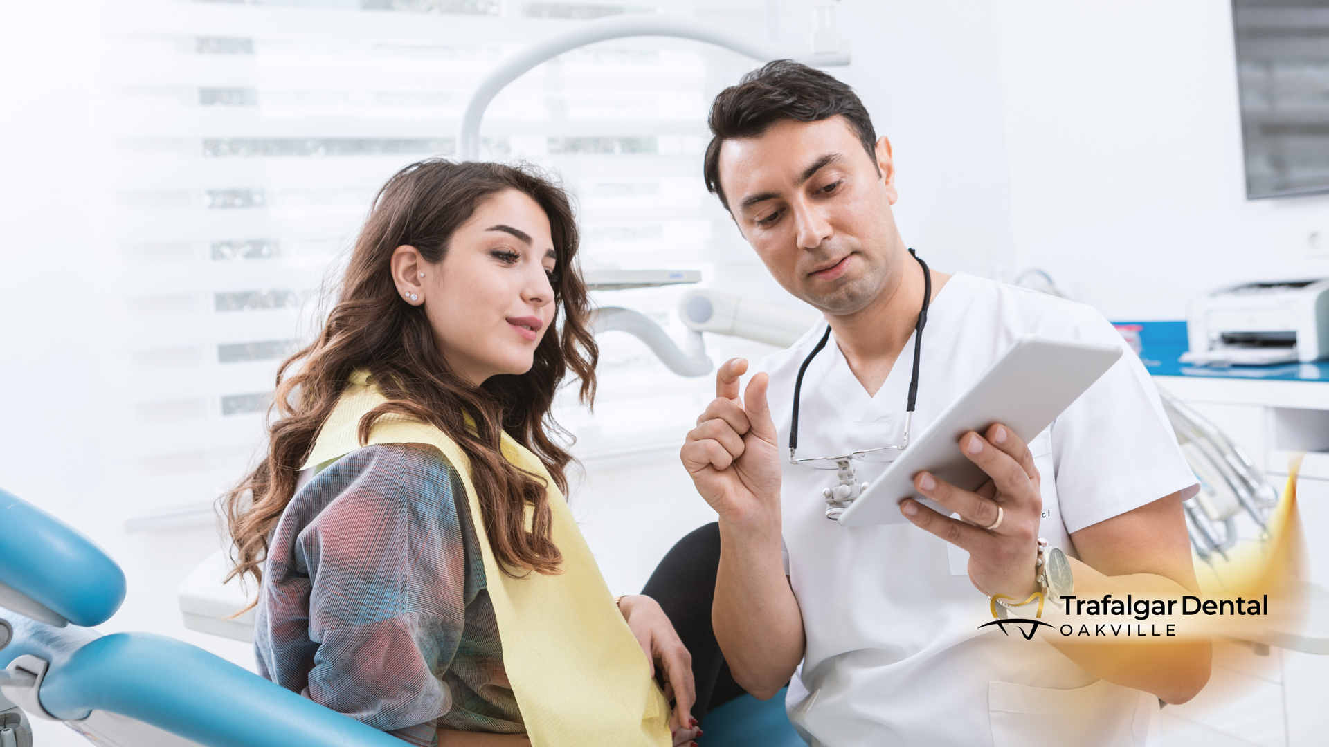 A dentist is talking to a patient in a dental chair while looking at a tablet.