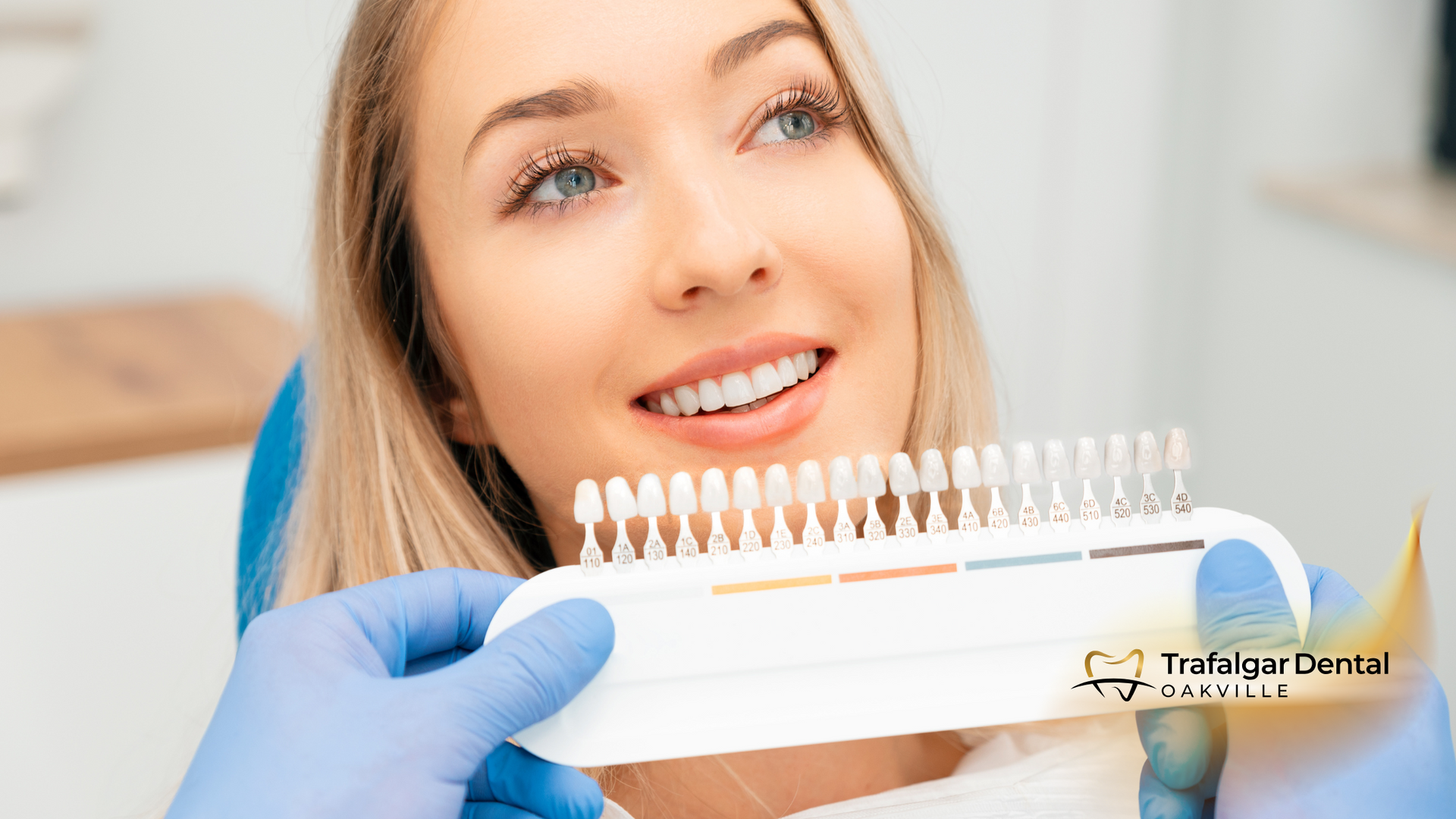 A woman is smiling while sitting in a dental chair while a dentist holds a tooth color chart.