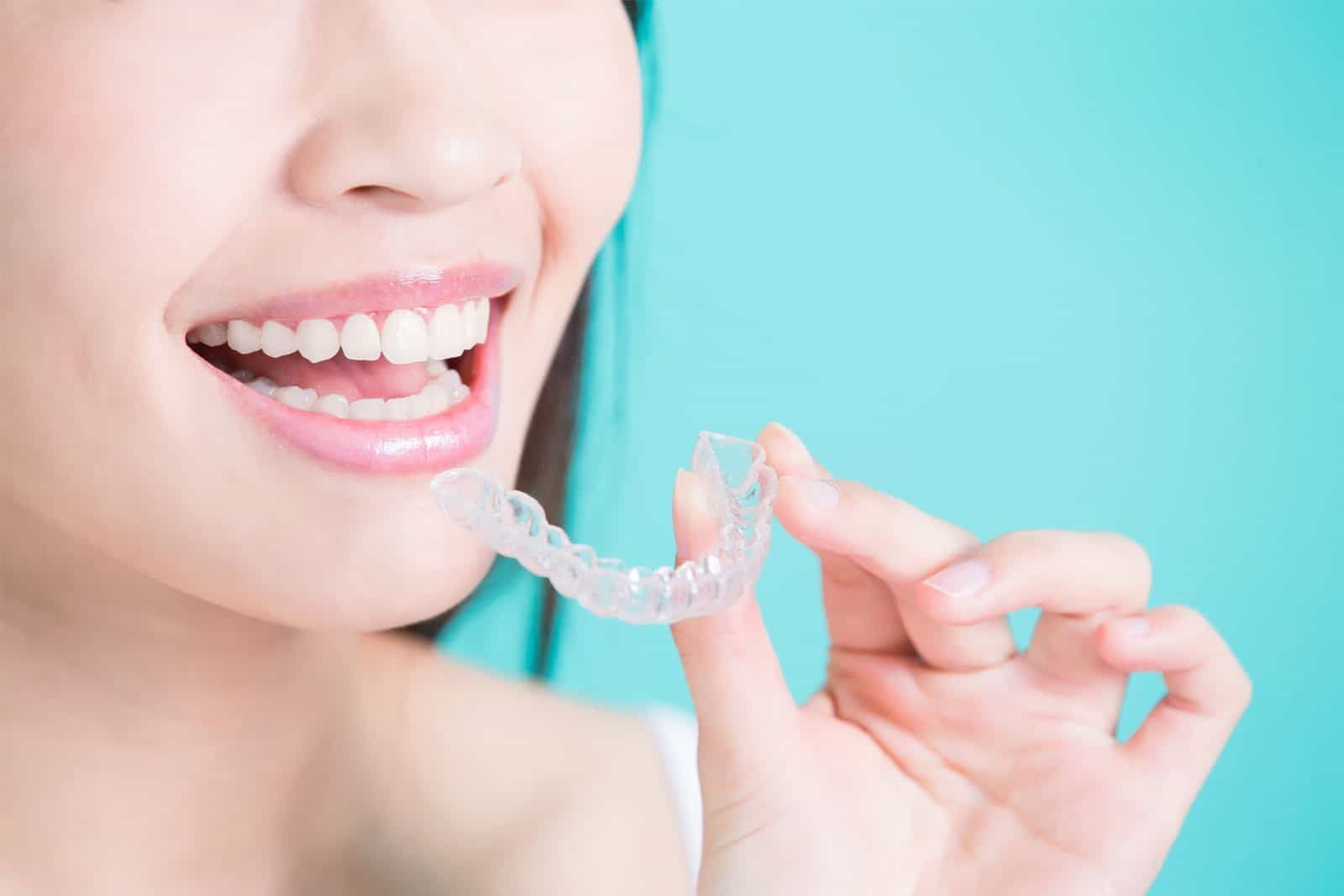 Woman smiling while holding a clear dental aligner near her mouth against a blue background