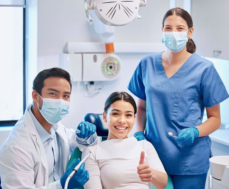 A woman is sitting in a dental chair with two dentists and a nurse.