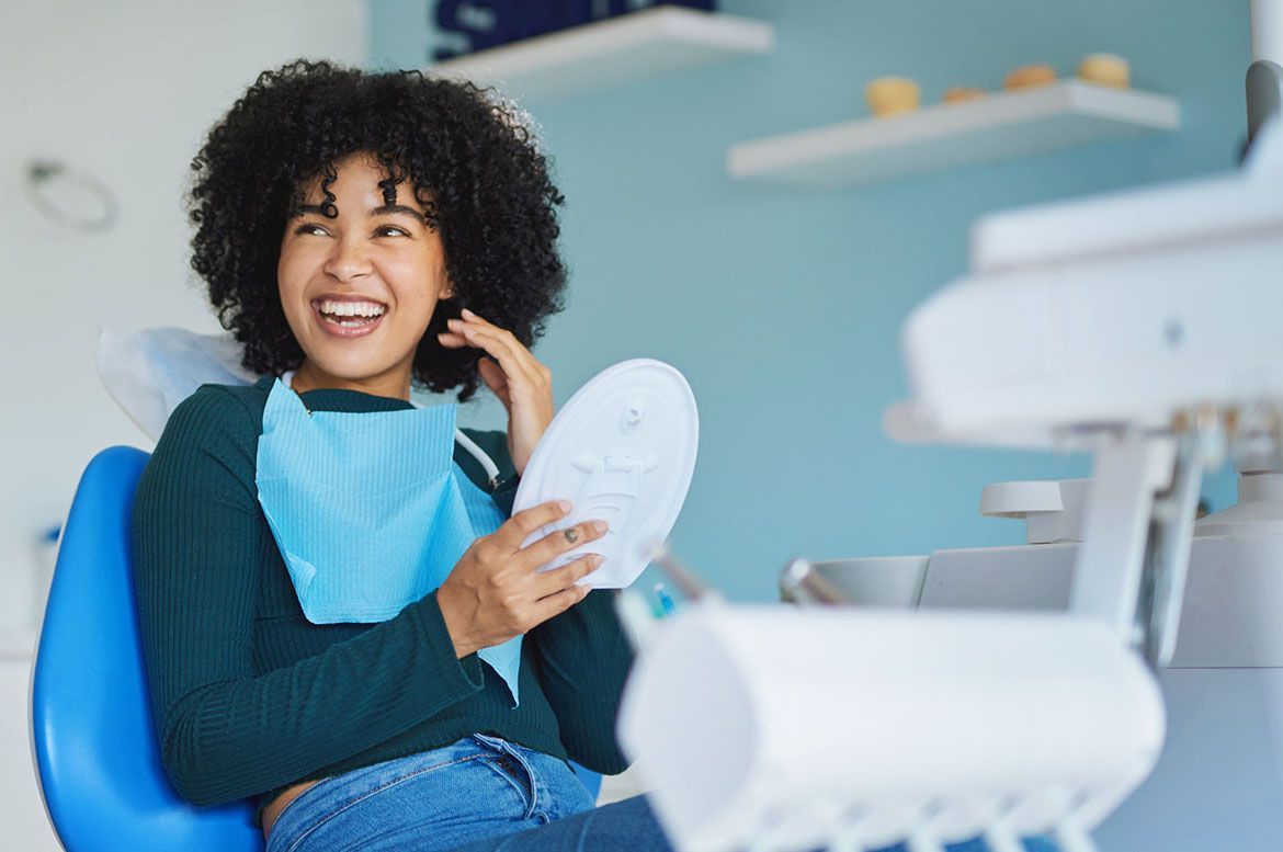 A woman is sitting in a dental chair looking at her teeth in a mirror.